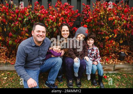A smiling family sit together on wooden log in front of red leaves Stock Photo