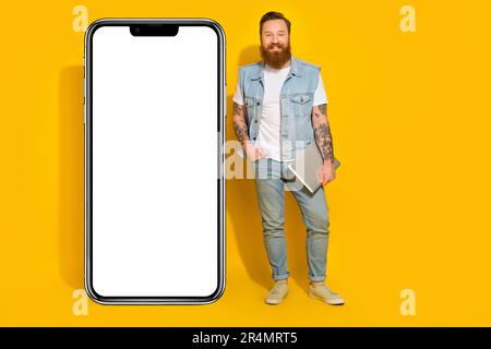 Photo of funny cute man wear jeans waistcoat holding modern device empty space isolated yellow color background Stock Photo
