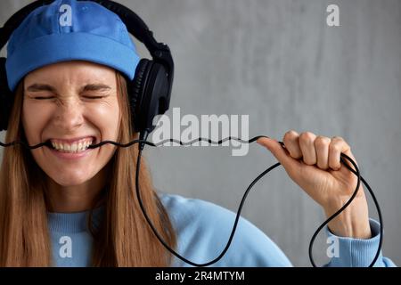 A young pretty long-haired DJ girl in a blue sweater and a funny hat wears headphones on her head and bites the wire. Studio shot, gray background Stock Photo