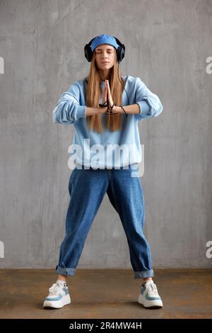 A young pretty long-haired DJ girl in a blue sweater, funny hat and headphones folded her hands in a prayerful meditation pose. Studio shot, gray back Stock Photo