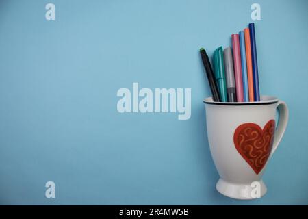 Heart-shaped mug photographed from above, placed on the edge of a blue background, with colorful pens inside. Stock Photo