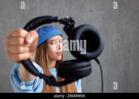 A young pretty long-haired DJ girl in a blue sweater and a funny blue hat holds black headphones in outstretched hands. Studio shot, gray background Stock Photo