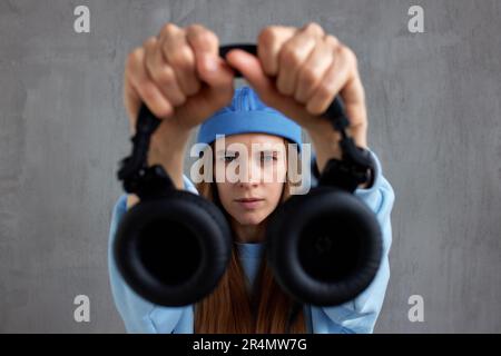 A young pretty long-haired DJ girl in a blue sweater and a funny blue hat holds black headphones in outstretched hands. Studio shot, gray background Stock Photo