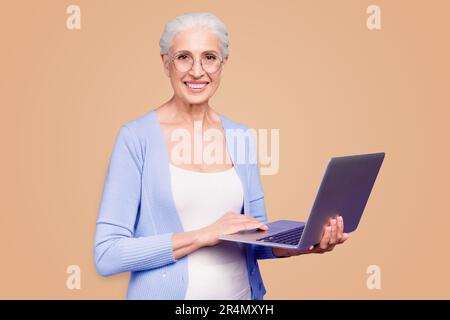 Grey haired old nice beautiful cheerful smiling woman wearing spectacles holding laptop learning online course. Isolated over violet purple background Stock Photo