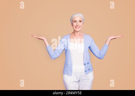 Beautiful nice cheerful smiling gray-haired old lady wearing casual, showing two options with palms. Isolated over pastel violet purple background Stock Photo