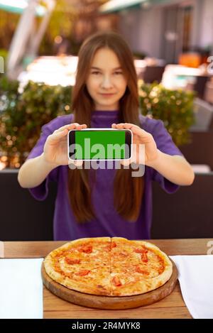 focus on phone in hands. young woman sitting in city cafe smiling showing smartphone blank green screen looking camera. client of pizzeria make order Stock Photo