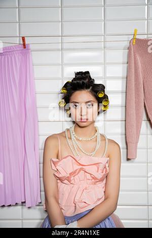 asian young woman with hair curlers standing in pink ruffled top and pearl necklace near clean and wet laundry hanging near white tiles, housework, ho Stock Photo