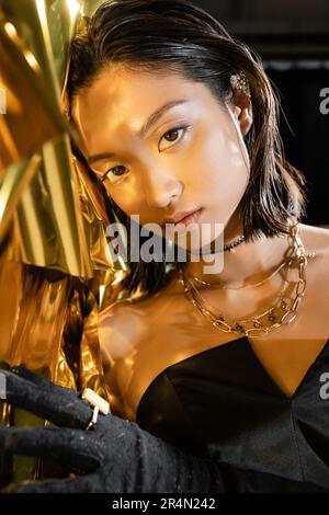 portrait of chic asian young woman with wet short hair posing in black strapless dress next to shiny background, model, looking at camera, wrinkled go Stock Photo