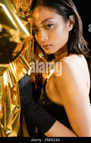 portrait of alluring asian young woman with wet short hair posing in strapless dress next to shiny background, model, looking at camera, wrinkled gold Stock Photo