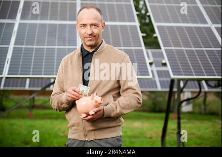 Investing in alternative energy innovations. Professional investor making investment in ecological resources. Young male in casual clothes holding piggy bank and smiling to camera. Stock Photo