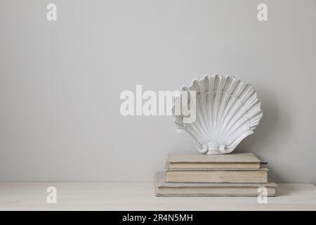 Stylish white vintage ceramic plate in shape of seashell on old books. Closeup of elegant wooden table. Empty beige wall background, nobody. Summer Stock Photo