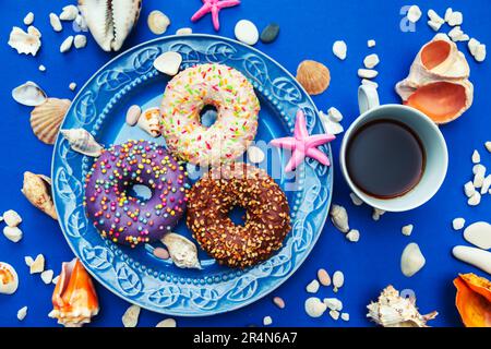 Assorted donuts in a plate on a blue background. Around them are symbols of the sea - shells, pebbles and stones. The concept of pleasure and relaxati Stock Photo
