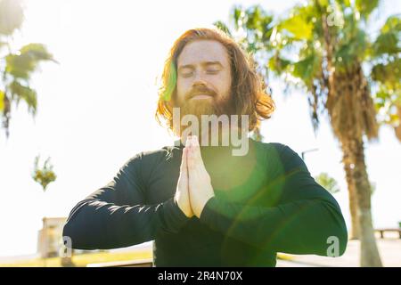 handsome redhaireb man with long beard wear black sporty suit doing yoga meditation in sunny beach Stock Photo