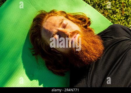 handsome redhaireb man with long beard wear black sporty suit doing yoga meditation in sunny beach Stock Photo