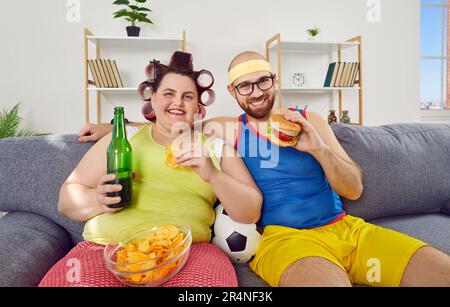 Funny couple watching football sport game sitting on sofa at home with beer and fast food. Stock Photo