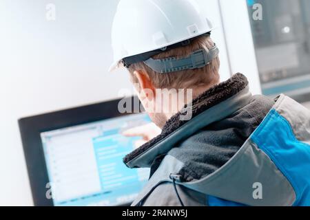 An engineer in a white helmet points to the touch screen of the natural gas automatic control system. Modern digital equipment in the gas industry. Back view. Authentic workflow. Stock Photo