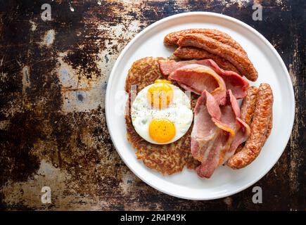 Indulge in a hearty breakfast feast: chipolata sausage, crispy bacon, and fried eggs atop golden hashbrowns, served on a pristine white plate Stock Photo