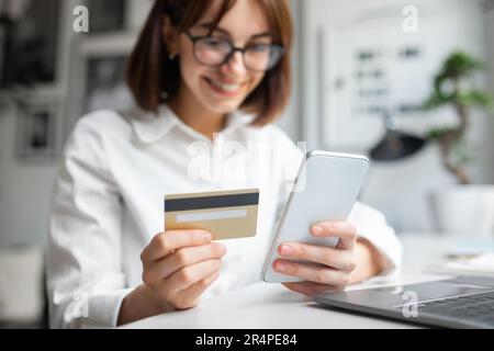 Young woman using smartphone and credit card, sitting at table with laptop, using modern mobile app for money trasfers Stock Photo