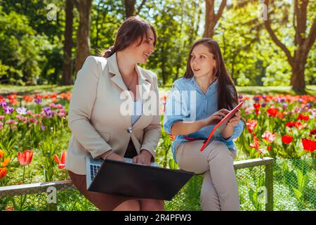 Two young businesswomen cowokers working in spring park using laptop and tablet. Remote work during break. Friends discussing tasks Stock Photo