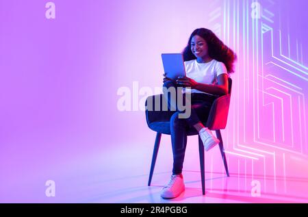 Online life. Young positive african american woman web surfing on digital tablet, sitting in armchair over neon light Stock Photo