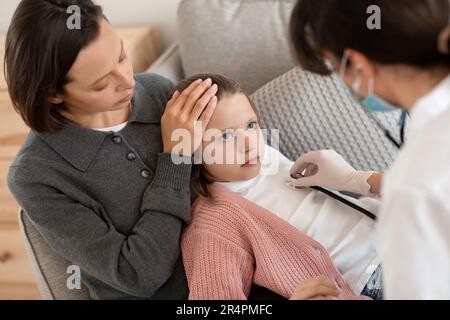 Worried sad caucasian middle aged mother touching forehead of little daughter, doctor listens to breathing Stock Photo