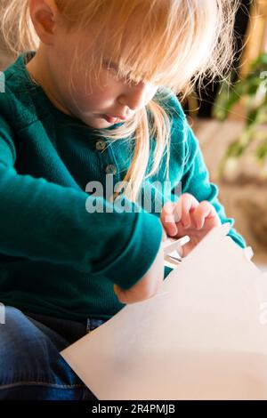 Four-year-old blonde Caucasian girl cutting paper with scissors. Stock Photo