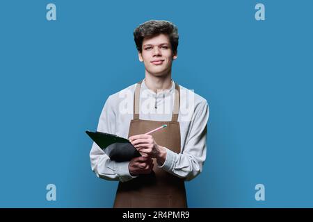 Young guy in apron making notes on clipboard, on blue studio background Stock Photo