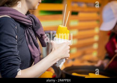 Cooked sweet corn on stick in hand of girl, selective focus. Summer vegan dinner or appetizer, grilling Stock Photo