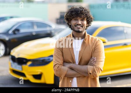 Portrait of a young smiling hispanic man against the background of a sports yellow car. He is standing on the cover, confident and smiling in front of the camera, with his arms crossed on his chest. Stock Photo