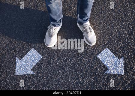 Choice of way. Man standing in front of drawn marks on road, closeup. White arrows pointing in different directions Stock Photo