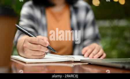 Cropped image of a young Asian female college student or a freelancer working on her tasks, writing and taking notes on her notebook at an outdoor caf Stock Photo