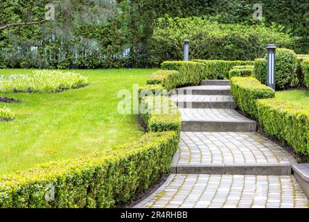 A winding paved step walkway in a beautiful park with decorative cut bushes and lanterns under soft sunlight. Copy space. Stock Photo
