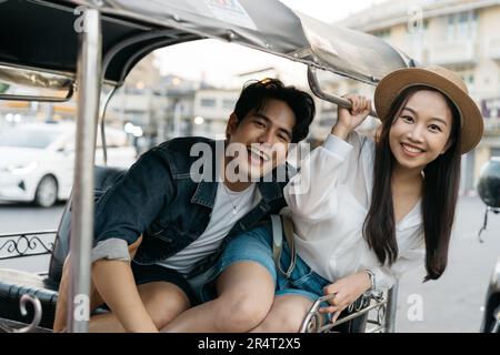 Happy and joyful Young Asian couple traveler tourists riding a tuk tuk tour, rickshaw style transportation on the street in Bangkok in Thailand - people traveling enjoying local culture concept Stock Photo