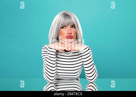 Dissatisfied young female model in gray wig in striped black and white shirt making face and looking away while leaning on hands at table against turq Stock Photo
