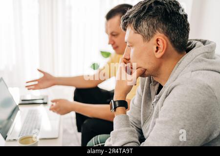 Two IT freelancer men typing in laptop working from home office. Two programmers sitting on couch doing code review. Distance learning online Stock Photo