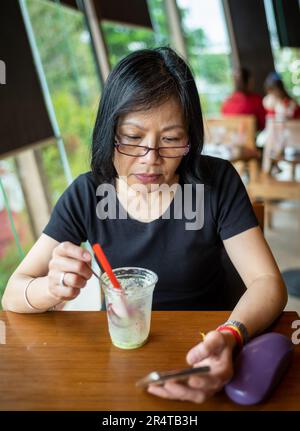 A middle aged Vietnamese woman finishes her coffee as she reads on her smartphone, Danang, Vietnam. Stock Photo