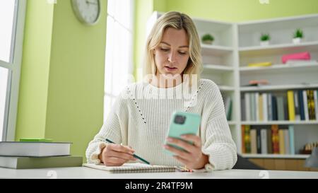 Young blonde woman student using smartphone taking notes at library university Stock Photo