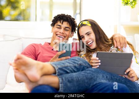 Cheerful biracial young couple using mobile phone and digital tablet while relaxing on sofa Stock Photo