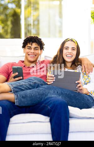 Portrait of happy biracial young couple using smartphone and digital tablet while relaxing on sofa Stock Photo