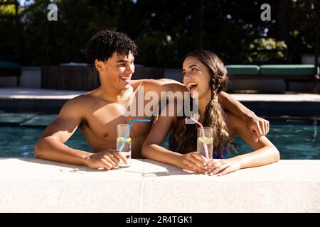 Happy biracial young couple holding drinks and talking in swimming pool during sunny day Stock Photo