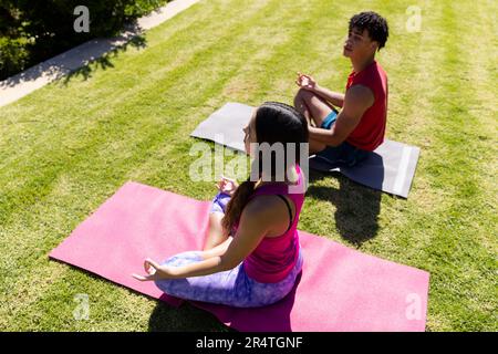High angle view of biracial young couple meditating while sitting on mats over field in yard Stock Photo
