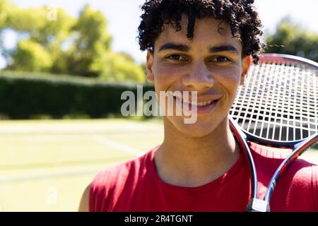 Close-up of biracial young man with tennis racket smiling in tennis court and looking at camera Stock Photo