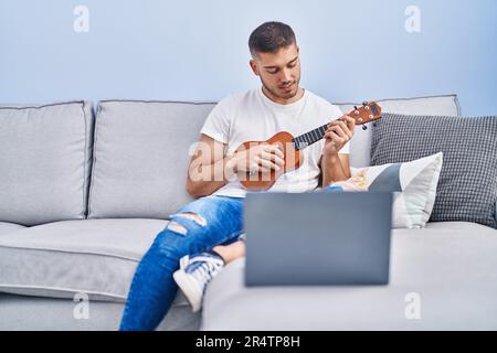 Young hispanic man having online ukelele class sitting on sofa at home Stock Photo
