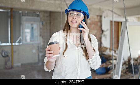 Young beautiful hispanic woman builder talking on smartphone drinking coffee at construction site Stock Photo