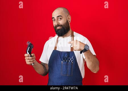 Young hispanic man with beard and tattoos wearing barber apron holding razor looking confident with smile on face, pointing oneself with fingers proud Stock Photo