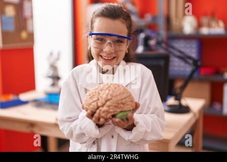 Little hispanic girl holding brain at science class at school smiling and laughing hard out loud because funny crazy joke. Stock Photo
