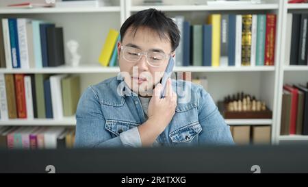 Young chinese man student using computer talking on smartphone at library university Stock Photo