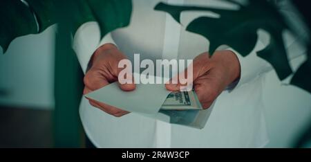 a man, wearing a white long-sleeve shirt, is putting, or taking out, a bunch of dollar notes in an envelope, in a panoramic format to use as web banne Stock Photo