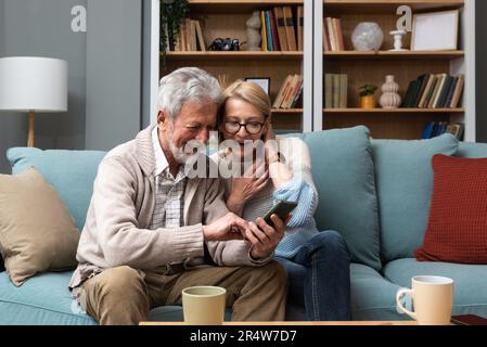 Relaxed mature 60s woman and 70s man older aged senior couple customer holding smartphone using mobile app texting message search ecommerce offers on Stock Photo