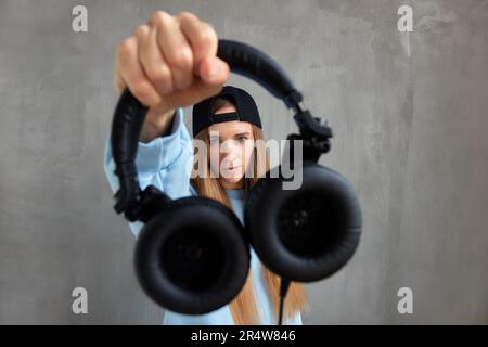 A young pretty long-haired DJ girl in a blue sweater and a funny blue hat holds black headphones in outstretched hands. Studio shot, gray background Stock Photo